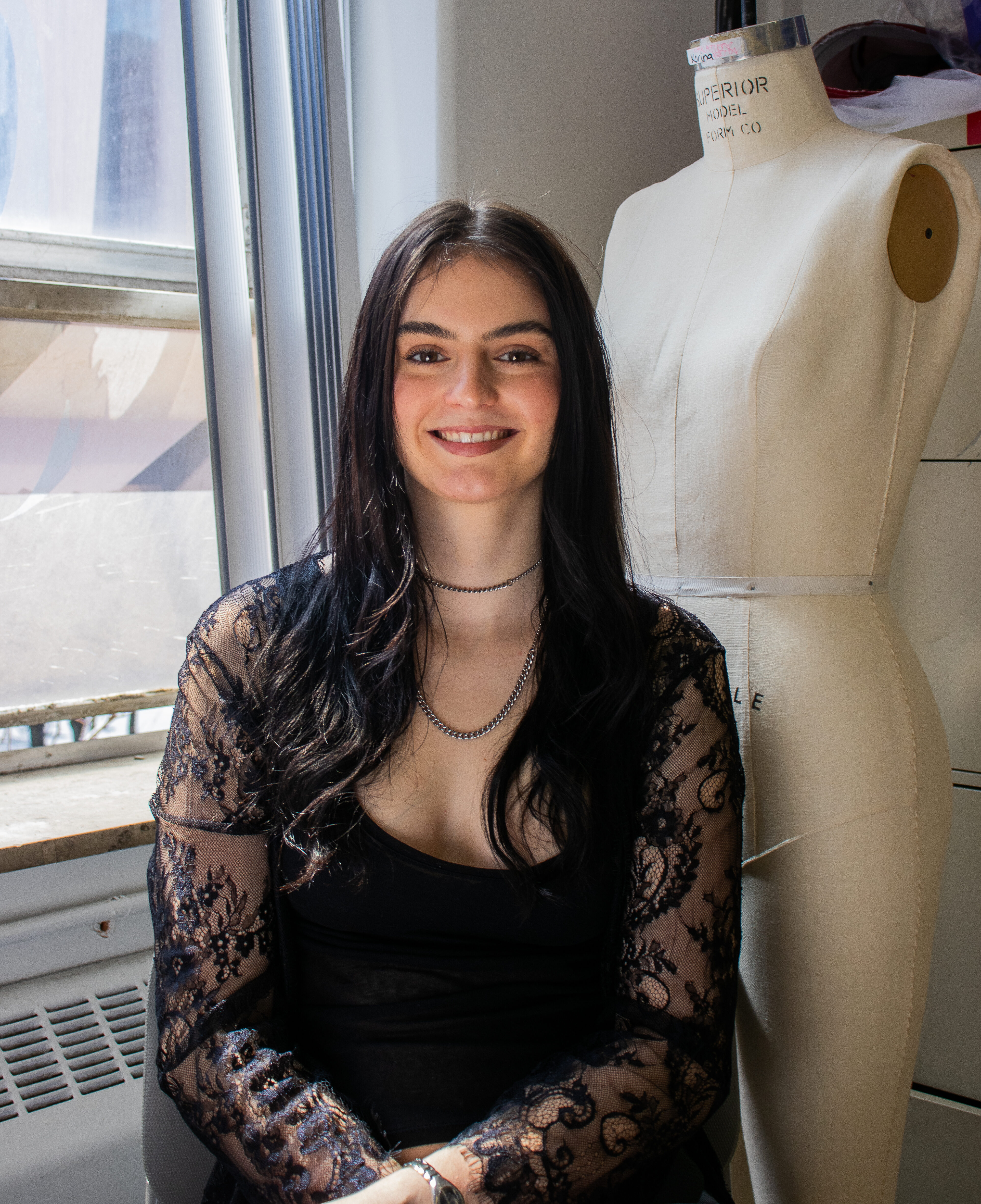 A young woman with long, dark, slightly wavy hair and a warm smile looks directly at the camera. She wears a black top with delicate lace sleeves and two silver chain necklaces of different lengths. She is seated between a brightly lit window on the left, showing a hint of outdoor architecture, and a cream-colored 'Superior Model Form Co' dress form on the right. The setting suggests a creative or fashion-related environment.