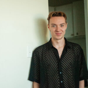 A fair-skinned young man with light brown hair and blue eyes smiles gently, looking directly at the viewer. He wears a sheer black short-sleeved button-up shirt with a textured chevron pattern and a thin gold chain. He is leaning against an off-white wall with a light switch on the left, and a blurred, darker room is visible in the background to his right.