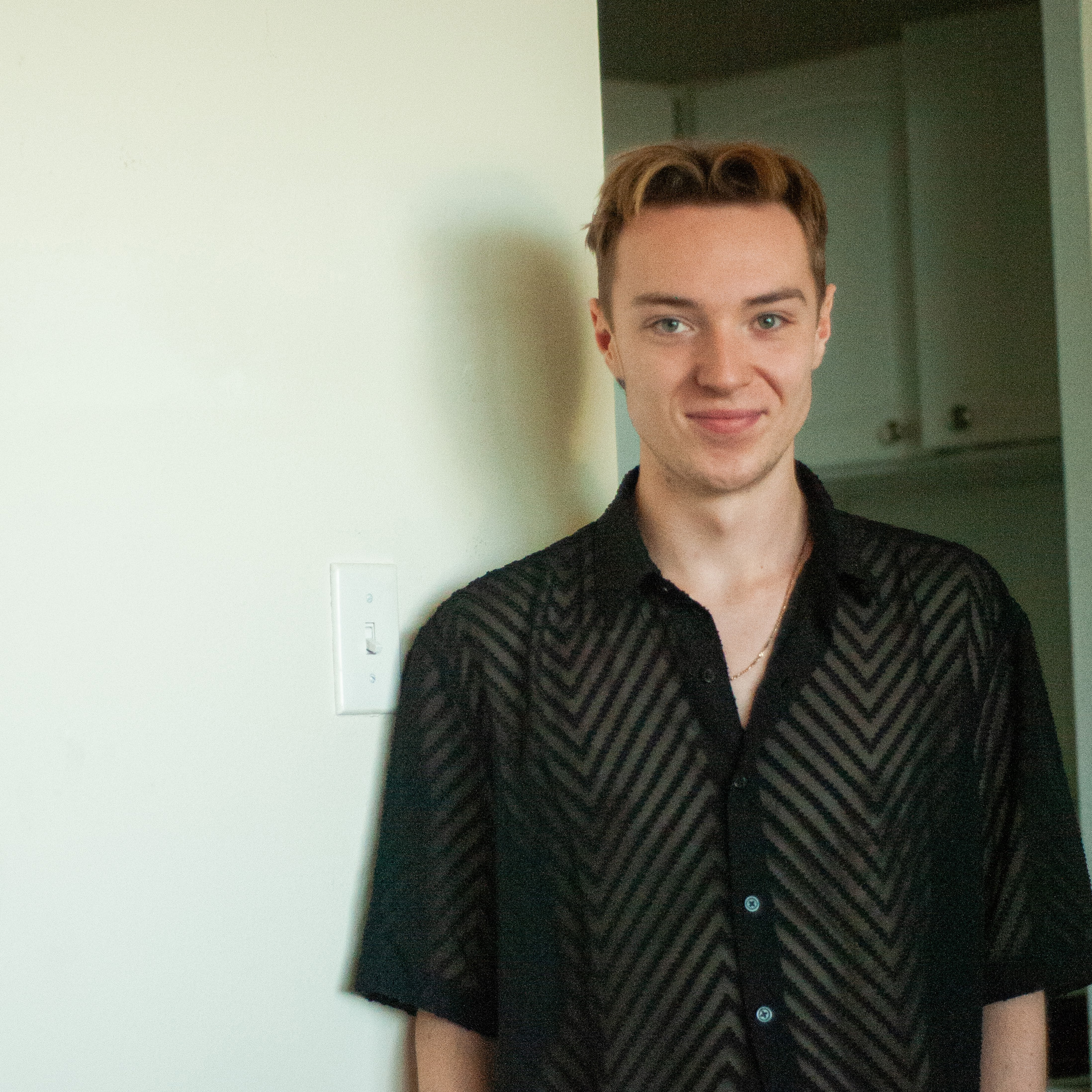 A fair-skinned young man with light brown hair and blue eyes smiles gently, looking directly at the viewer. He wears a sheer black short-sleeved button-up shirt with a textured chevron pattern and a thin gold chain. He is leaning against an off-white wall with a light switch on the left, and a blurred, darker room is visible in the background to his right.
