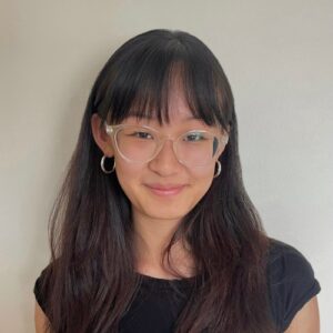 A young East Asian woman with long dark hair and bangs smiles gently while wearing clear-framed glasses and silver hoop earrings. She is wearing a black t-shirt against a plain light-colored background.